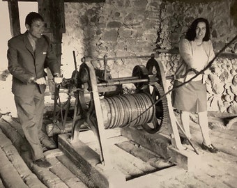 Könnte beinhalten: Ein Schwarzweißfoto von zwei Personen, die eine große, industrielle Winde bedienen. Die Winde hat eine große, metallene Trommel, um die ein dickes Seil gewickelt ist. Die Personen stehen auf einer Holzplattform.