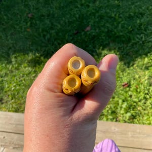 May include: Three small, yellow, cylindrical objects with a hole in the center are held in a hand. The objects have a textured, possibly carved, design. The background is a blurred view of green grass.
