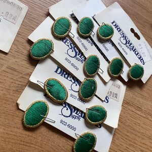 May include: A pack of green oval-shaped fabric buttons with gold trim and a gold pin on the back. The buttons are on a white card with the text "Dressmakers Supply" and a phone number.