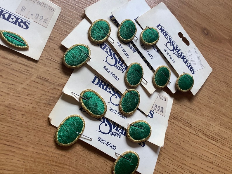 May include: A pack of green oval-shaped fabric buttons with gold trim and a gold pin on the back. The buttons are on a white card with the text "Dressmakers Supply" and a phone number.