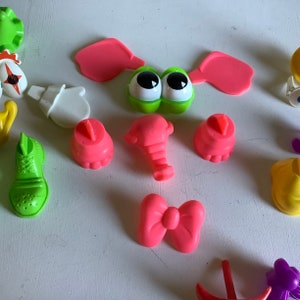 Może przedstawiać: Zestaw kolorowych plastikowych element&oacute;w do gry dla dzieci. Elementy obejmują zieloną twarz z oczami, r&oacute;żowe uszy, r&oacute;żowy trąbę, r&oacute;żowe stopy, r&oacute;żową kokardkę, zielony but, białą rękawiczkę i ż&oacute;łtą koronę.