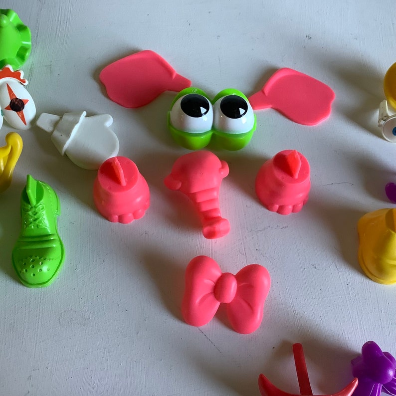 Może przedstawiać: Zestaw kolorowych plastikowych element&oacute;w do gry dla dzieci. Elementy obejmują zieloną twarz z oczami, r&oacute;żowe uszy, r&oacute;żowy trąbę, r&oacute;żowe stopy, r&oacute;żową kokardkę, zielony but, białą rękawiczkę i ż&oacute;łtą koronę.