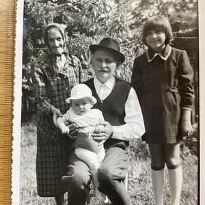 Peut inclure: Photographie en noir et blanc d'une famille posant à l'extérieur. Une femme âgée en manteau à motifs et foulard se tient à côté d'un homme chapeauté et vêtu d'un gilet tenant un bébé. Une jeune fille en robe est à proximité.