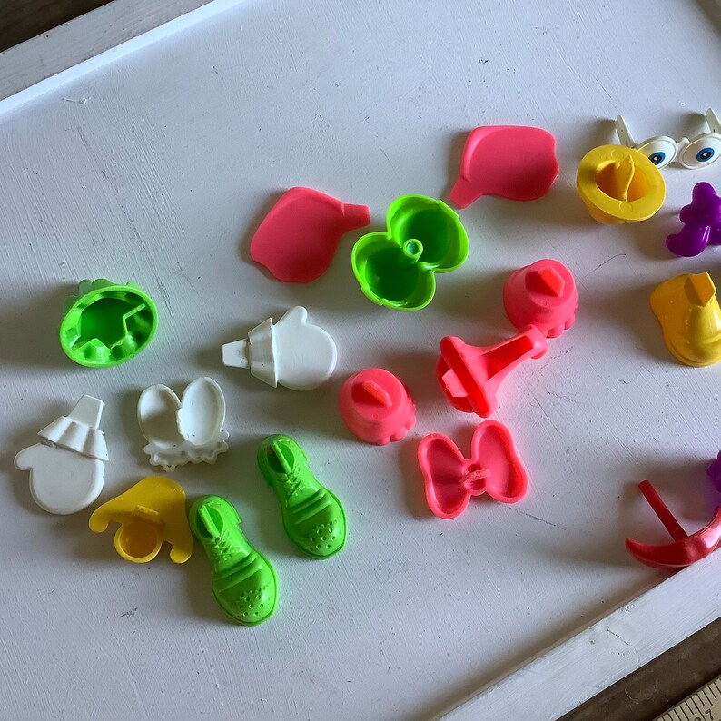Może przedstawiać: Zbi&oacute;r kolorowych plastikowych form do zabawkowego jedzenia, w tym para but&oacute;w, kokarda, kwiat, kapelusz i para rękawiczek. Formy są przeznaczone do użytku z plasteliną lub masą modelarską.