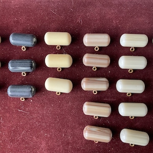 Puede incluir: Surtido de 18 botones de plástico blanco, negro y marrón con un agujero en el centro. Los botones están dispuestos en filas sobre una superficie roja.