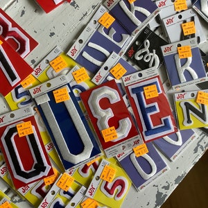 May include: A collection of felt letter patches in various colours, including red, blue, white, and black. The patches are arranged on a white surface and have price tags attached. The letters include Q, U, E, N, and N.