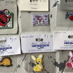 May include: An assortment of iron-on patches featuring various designs, including Santa Claus, a snowman, an aeroplane, a hedgehog, a bunny, and a pink bunny. The patches are packaged on white cards with the text "MCI Dressmaker Supplies" and phone number (416) 922-6000.