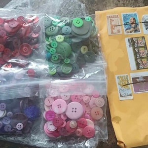 May include: Three bags of assorted buttons in various colors, including red, green, purple, and pink. The bags are sealed and ready to be shipped.