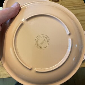 K&ouml;nnte beinhalten: Ein rosa Keramikteller mit einem erh&ouml;hten Rand und einem kreisf&ouml;rmigen Design in der Mitte. Auf der Unterseite des Tellers steht "Manufactured by MAPLEX 1977" gedruckt.