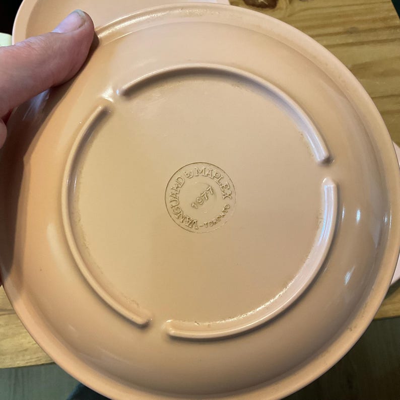 K&ouml;nnte beinhalten: Ein rosa Keramikteller mit einem erh&ouml;hten Rand und einem kreisf&ouml;rmigen Design in der Mitte. Auf der Unterseite des Tellers steht "Manufactured by MAPLEX 1977" gedruckt.