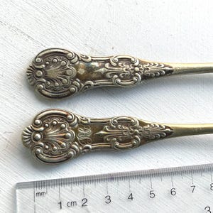 Op de afbeelding: Twee zilveren serveerslepels met sierlijke handvatten. De handvatten hebben een bloemmotief met een verhoogd middelste deel. De lepels zijn 2 cm lang.