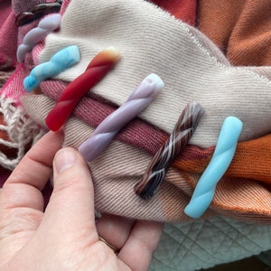 Op de afbeelding: Vijf kleurrijke gedraaide haarclips in verschillende kleuren, waaronder rood, paars, bruin en blauw, worden getoond op een beige en oranje gestreepte stof.