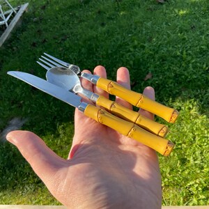 May include: A set of four pieces of silverware: a fork, spoon, and knife. The silverware has yellow bamboo-style handles and silver metal heads. The items are held in a hand against a green grass background.