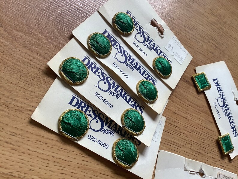May include: Six green and gold fabric covered buttons on white cards with the text "Dressmakers Supply" and the phone number 922-6000. The price is $1.99.