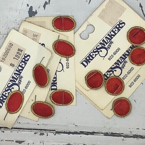 May include: A close-up of red oval fabric patches with gold trim and a single pin attached to each. The patches are on white card stock with the text "Dressmakers Supply" and a phone number.