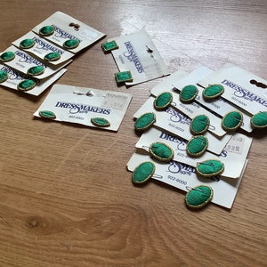 May include: A collection of green and gold oval-shaped buttons with the text "Dressmakers Supply" on the packaging. The buttons are on white cards with the text "922-8000" printed on them.
