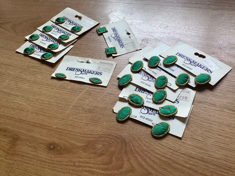 May include: A collection of green and gold oval-shaped buttons with the text "Dressmakers Supply" on the packaging. The buttons are on white cards with the text "922-8000" printed on them.