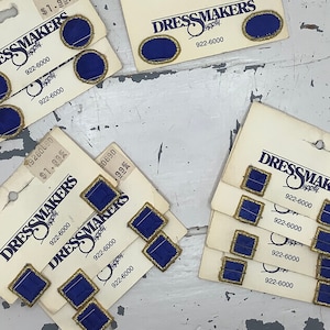 May include: Assortment of cards with blue and gold trim featuring the text "Dressmakers Supply" and the phone number 922-6000. The cards are of various sizes and shapes, and some feature small blue and gold buttons.
