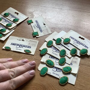 May include: Assortment of green oval-shaped buttons with gold trim on white cards with the text "Dressmakers Supply" and phone number "922-6000".