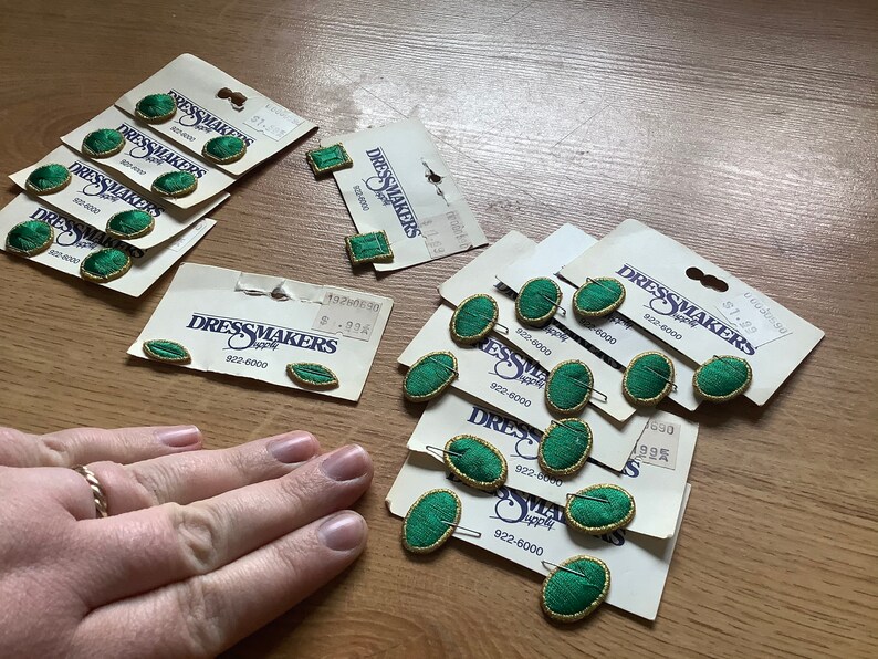 May include: Assortment of green oval-shaped buttons with gold trim on white cards with the text "Dressmakers Supply" and phone number "922-6000".