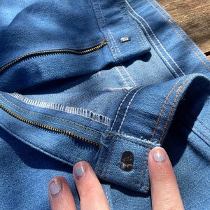 Op de afbeelding: Close-up van een paar blauwe denim jeans met een rits en een knoopsluiting. De jeans heeft witte stiksels en een bruine naad.