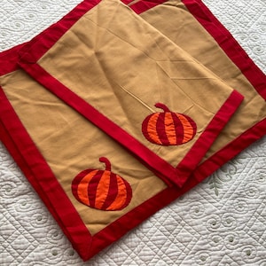 May include: Two tan placemats with red trim and a pumpkin applique in orange and red.