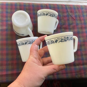 Puede incluir: Cuatro tazas de cerámica blanca con un diseño floral azul alrededor del borde. Una taza está boca abajo, revelando el nombre de la marca. Las tazas tienen un asa y una forma ligeramente acampanada. Las tazas están sobre una superficie a cuadros rojos y azules.