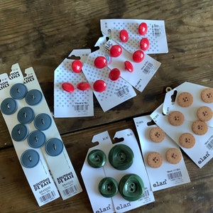 Può includere: Assortimento di bottoni in vari colori e dimensioni, tra cui rosso, blu, verde e marrone. I bottoni sono confezionati su carte con il marchio "elan" e "bottoni base".