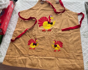 May include: A tan apron with red ties and three embroidered turkeys. The turkeys are yellow with red wings and black bow ties.