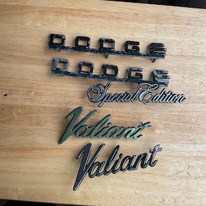 Può includere: Diversi emblemi di auto d'epoca sono esposti su una superficie di legno. Due emblemi neri "Dodge" sono in alto. Sotto ci sono un emblema "Special Edition" e due emblemi "Valiant", uno verde e uno nero.