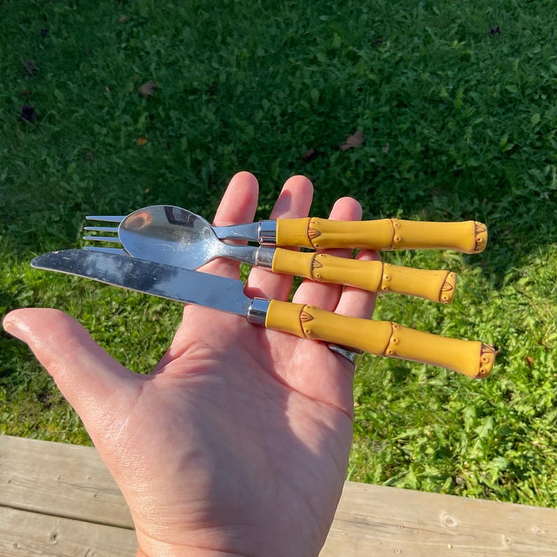 May include: A stainless steel fork, spoon, and knife set with bamboo-style yellow handles. The flatware is held in a hand against a green grass background. The knife blade is approximately 8 inches long.