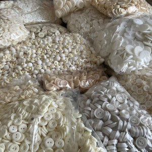 May include: A selection of white and cream buttons in various sizes and shapes, packaged in clear plastic bags. The buttons are arranged in a random pattern, creating a visually appealing display.