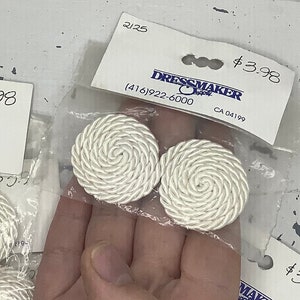 Peut inclure: Deux boutons ronds blancs en forme de corde avec un motif en spirale. Les boutons sont emballés sur une carte blanche avec le texte "Dressmaker Supply" et une étiquette de prix de 3,98 $.