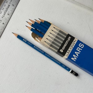Op de afbeelding: Een doos blauwe STAEDTLER MARS-LUMOGRAPH potloden met de tekst "100 3H" en een enkel potlood ernaast. De potloden zijn geslepen en klaar voor gebruik. De doos is blauw met witte tekst.