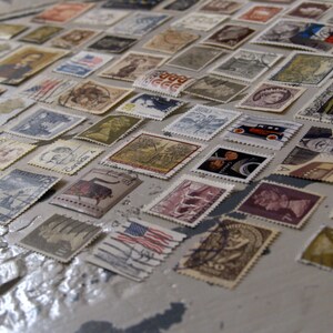 May include: A collection of vintage postage stamps featuring a variety of designs and subjects, including portraits, landscapes, and transport. The stamps are arranged in rows on a light grey surface.