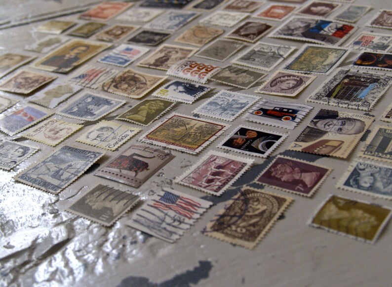 May include: A collection of vintage postage stamps featuring a variety of designs and subjects, including portraits, landscapes, and transport. The stamps are arranged in rows on a light grey surface.