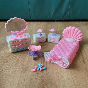 Può includere: Un set da camera da letto per bambole rosa e bianco con un letto a forma di conchiglia, due comodini, un comò con specchio e un tavolino con piano rosa. Il set include diversi piccoli accessori a forma di conchiglia.