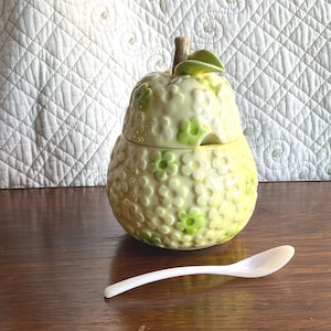 May include: A ceramic pear-shaped container with a lid, decorated with small green and yellow flowers. A small white spoon rests on a wooden surface in front of the container. The container has a stem and leaf detail.