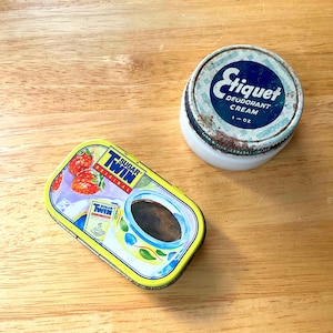 May include: A vintage tin of Sugar Twin original sweetener with strawberries and a cup of coffee. Next to it is a white jar of Etiquet Deodorant Cream, 1 oz. Both items are on a wooden surface.
