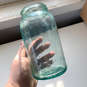 Puede incluir: Un tarro de vidrio transparente con un tono verde. El tarro tiene una boca ancha y está estampado con las palabras "Improved Mason" y un diseño de corona.