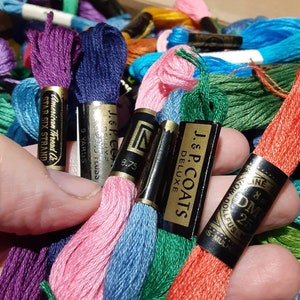 May include: A close-up of a hand holding a variety of embroidery floss spools in different colors, including purple, blue, pink, green, and orange. The spools are labeled with the brand name "J & P. Coats Deluxe" and "American Thread Co. Star Six Strand".