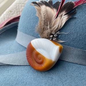 Puede incluir: Un broche de color marrón y blanco con forma de concha con un acento de pluma. El broche está en un sombrero azul.