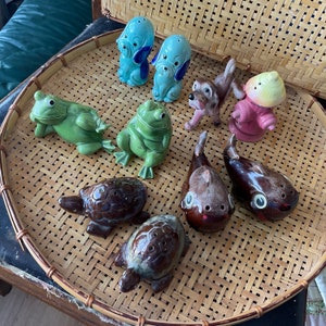 May include: A collection of ceramic animal figurines, including frogs, dogs, and turtles, in various colours, such as blue, green, brown, and white. The figurines are arranged on a woven wicker tray.