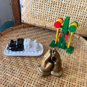 Puede incluir: Una colección de saleros y pimenteros decorativos y un juguete. Los saleros y pimenteros incluyen un juego de trenes en blanco y negro y un par de manos doradas. También hay un juguete verde y amarillo.