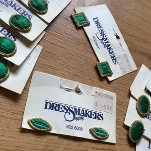 May include: Assortment of green and gold fabric covered buttons with a Dressmakers Supply label. The buttons are in various shapes, including round, oval, and square. The label includes the company name, phone number, and price of the buttons.