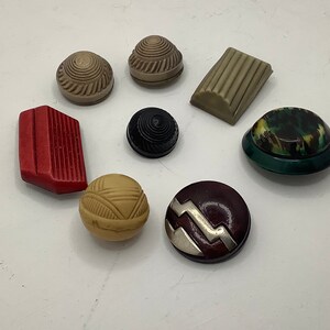 Op de afbeelding: Assortiment van acht decoratieve knopen in verschillende kleuren en patronen, waaronder rood, bruin, zwart, wit en groen. De knopen zijn gemaakt van verschillende materialen, zoals plastic, metaal en hout.