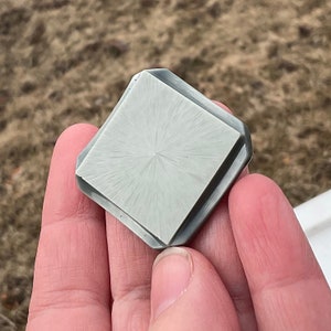 May include: A square-shaped, gray, ceramic tile with a textured surface. The tile has a radial pattern of lines radiating from the center.