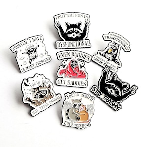 May include: A selection of enamel pins with raccoon designs and humorous slogans. The pins are in black, white, and red, with phrases such as "Dysfunctional" and "Stay Trashy." The pins are ideal for jackets or bags.