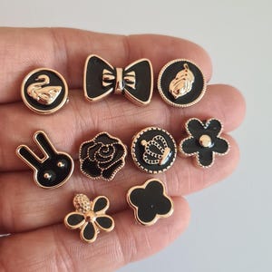 Op de afbeelding: Een verzameling zwarte en gouden decoratieve knopen in verschillende vormen, waaronder een zwaan, strik, blad, konijn, roos, kroon en bloemen. De knopen hebben een glanzende zwarte emaille afwerking met goudkleurige metalen accenten.