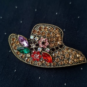 May include: A hat-shaped brooch featuring a gold-tone metal base encrusted with numerous amber-colored rhinestones. The design includes decorative floral accents made of red, pink, and dark brown crystals, along with marquise-cut purple and green stones.
